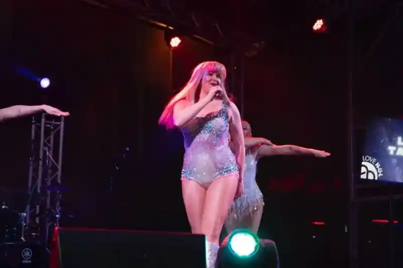 A performer in a sparkling silver outfit sings passionately on stage, with colorful lights illuminating the scene. Two backing dancers are positioned to the side, striking poses and enhancing the energetic atmosphere of the performance.