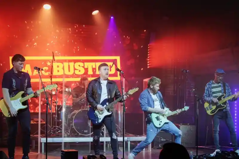 A band performs on stage with colorful lights and a neon sign in the background. The lead guitarist strikes a dynamic pose while the other members play electric guitars and drums.