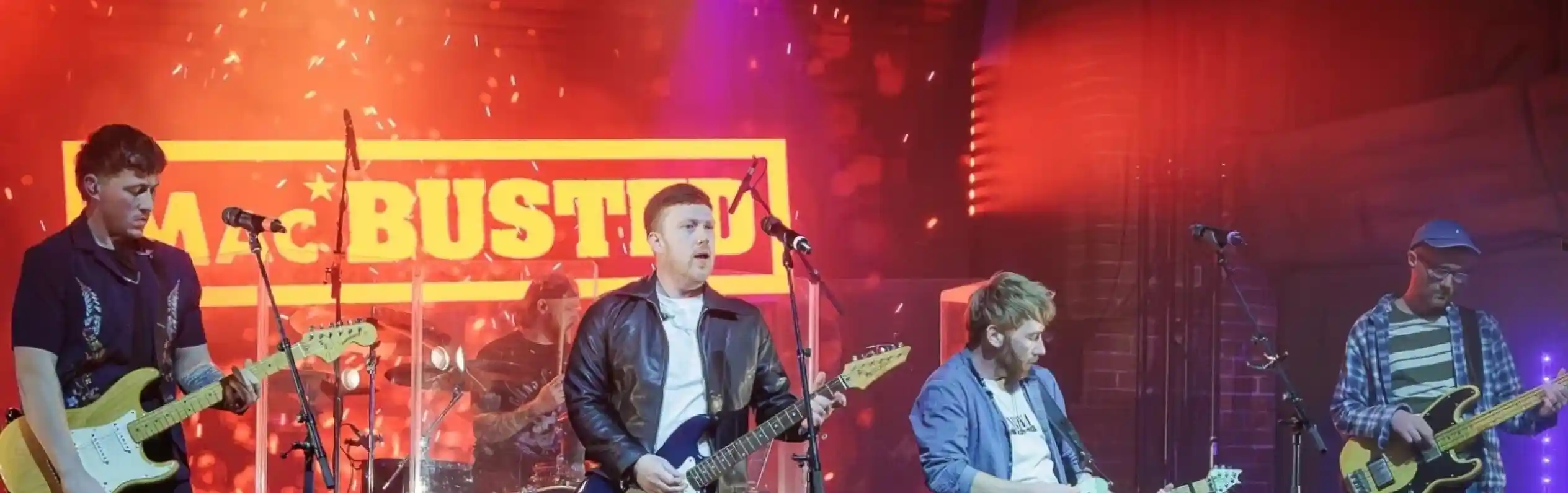 A band performs on stage with colorful lights and a neon sign in the background. The lead guitarist strikes a dynamic pose while the other members play electric guitars and drums.