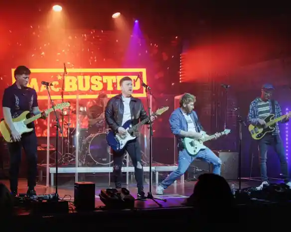 A band performs on stage with colorful lights and a neon sign in the background. The lead guitarist strikes a dynamic pose while the other members play electric guitars and drums.