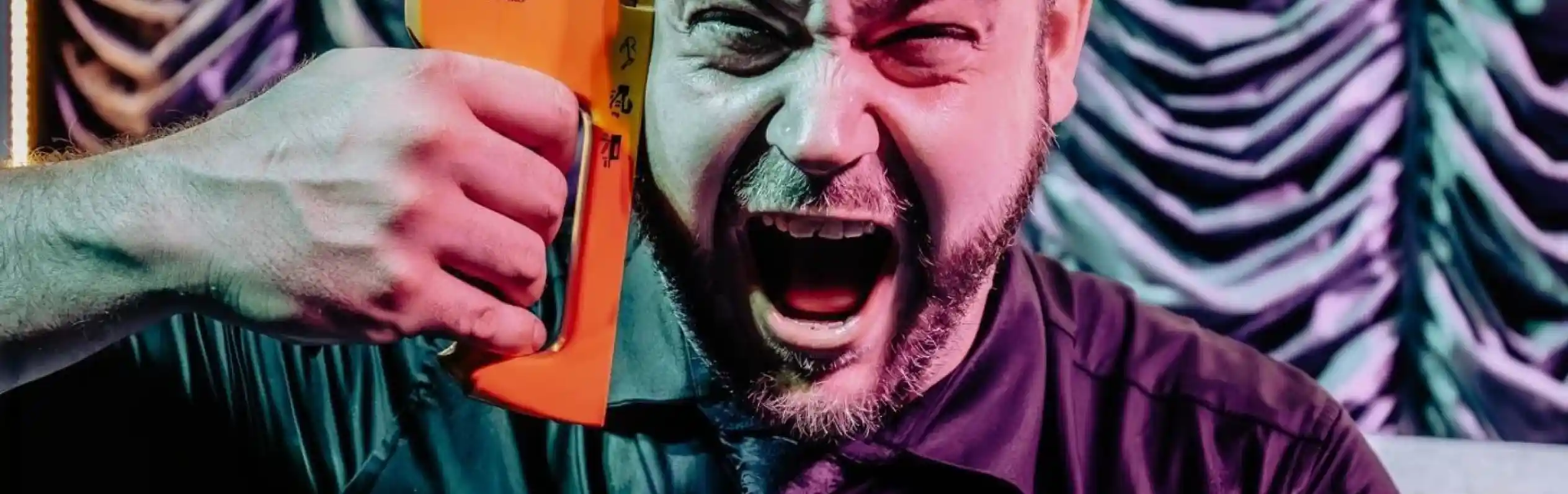 A frustrated man in a black shirt and tie holds an orange stapler to his head, yelling in a dramatic expression against a theatrical backdrop.