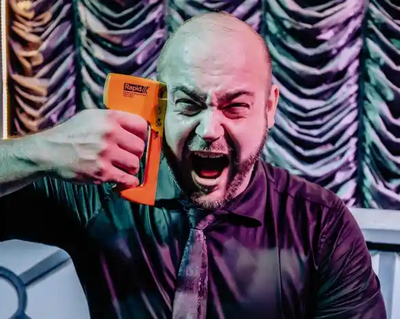 A frustrated man in a black shirt and tie holds an orange stapler to his head, yelling in a dramatic expression against a theatrical backdrop.