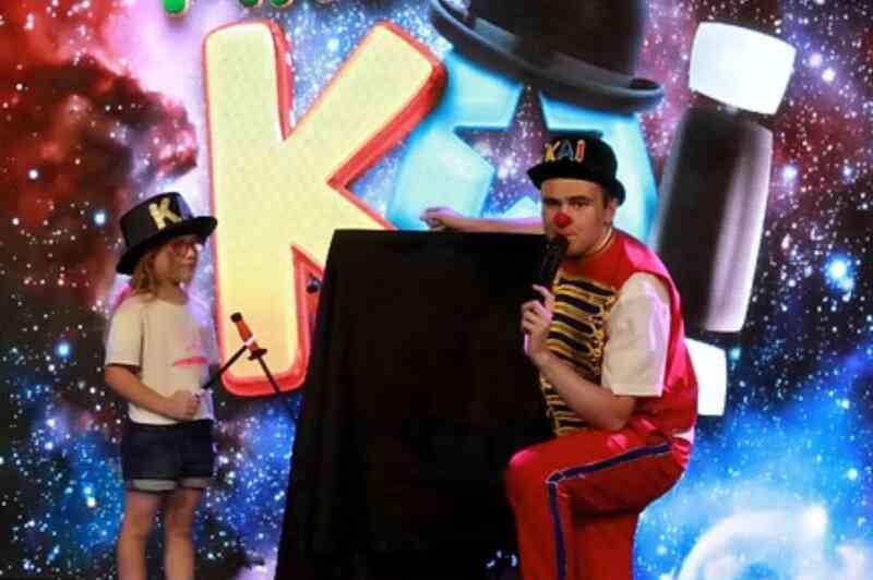 A performer in a colorful outfit kneels beside a child holding a toy microphone. Both are in front of a vibrant, cosmic-themed backdrop with playful letters. The child wears a white shirt and denim shorts, while the performer is in red and blue attire, with a hat that says "KAI."