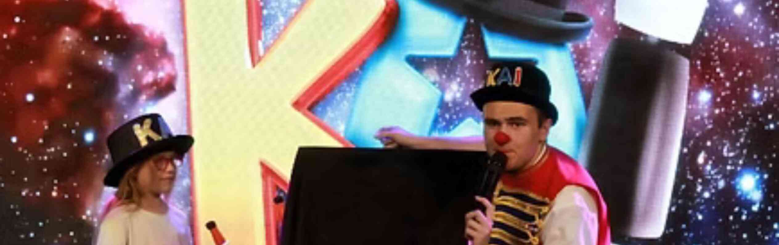 A performer in a colorful outfit kneels beside a child holding a toy microphone. Both are in front of a vibrant, cosmic-themed backdrop with playful letters. The child wears a white shirt and denim shorts, while the performer is in red and blue attire, with a hat that says "KAI."
