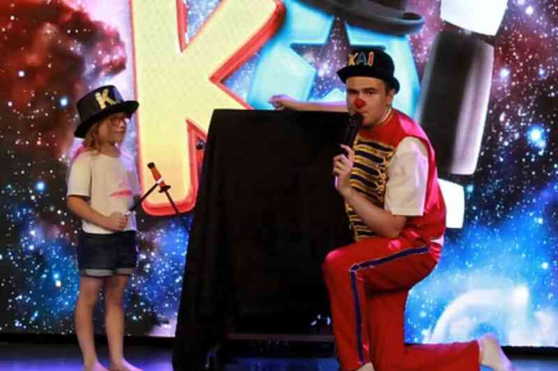 A performer in a colorful outfit kneels beside a child holding a toy microphone. Both are in front of a vibrant, cosmic-themed backdrop with playful letters. The child wears a white shirt and denim shorts, while the performer is in red and blue attire, with a hat that says "KAI."