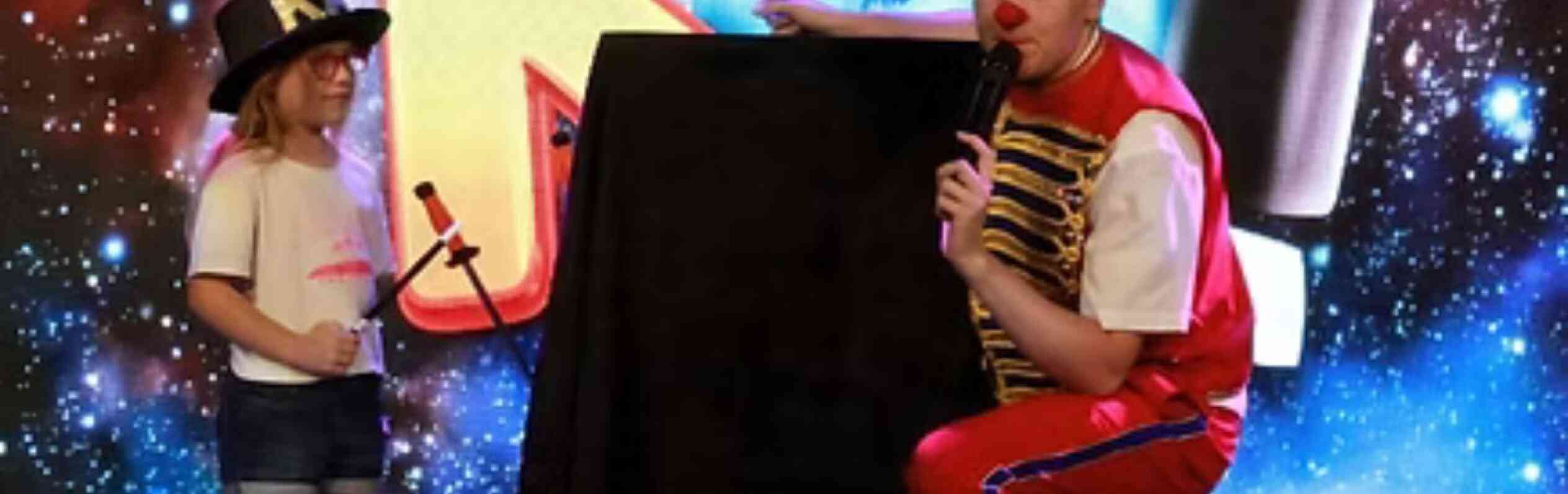 A performer in a colorful outfit kneels beside a child holding a toy microphone. Both are in front of a vibrant, cosmic-themed backdrop with playful letters. The child wears a white shirt and denim shorts, while the performer is in red and blue attire, with a hat that says "KAI."