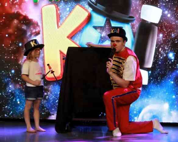 A performer in a colorful outfit kneels beside a child holding a toy microphone. Both are in front of a vibrant, cosmic-themed backdrop with playful letters. The child wears a white shirt and denim shorts, while the performer is in red and blue attire, with a hat that says "KAI."