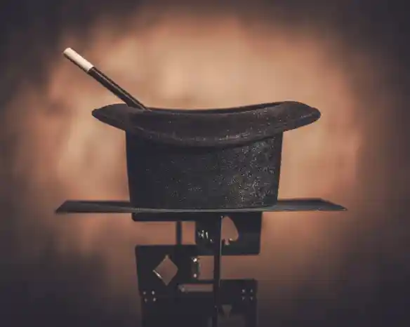 A black magician's hat sits on a stand, with a white magic wand resting inside it. The background is softly blurred, creating a warm ambiance.