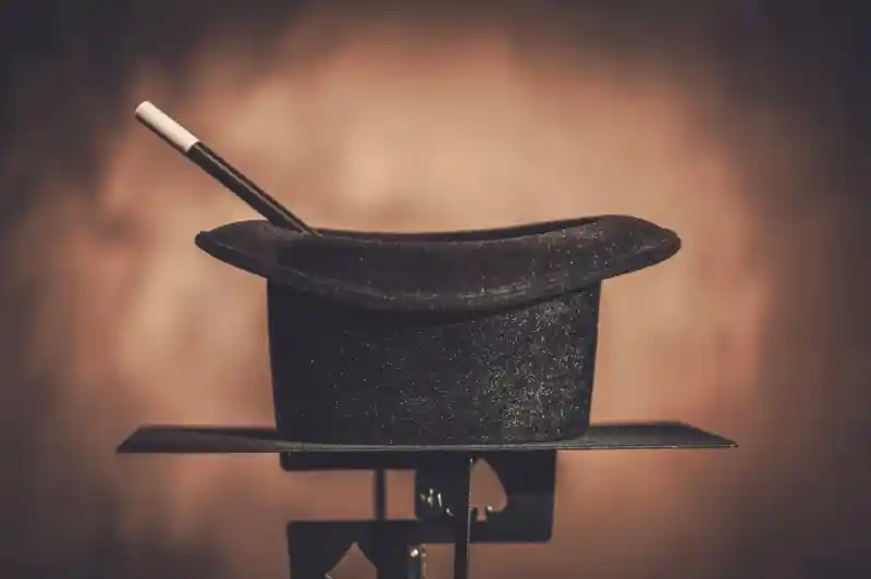 A black magician's hat sits on a stand, with a white magic wand resting inside it. The background is softly blurred, creating a warm ambiance.