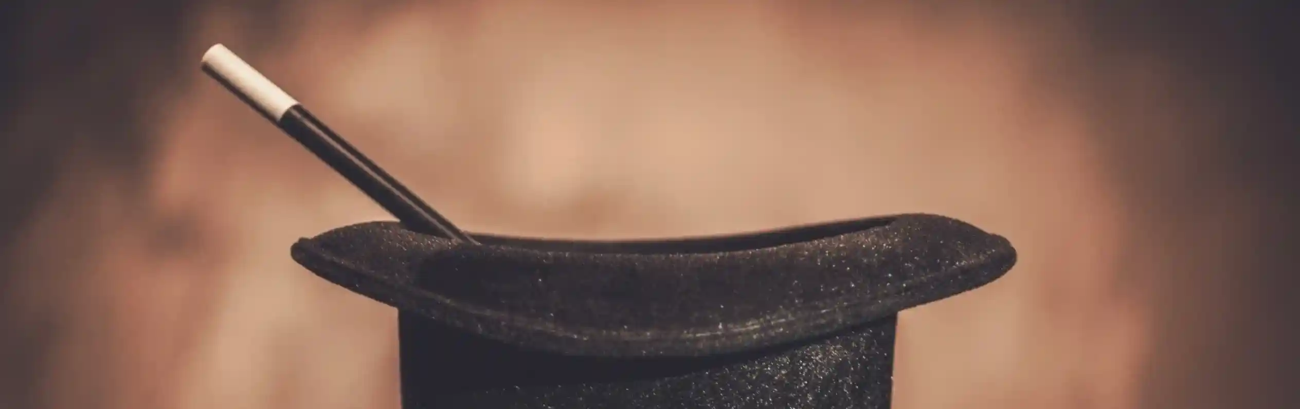 A black magician's hat sits on a stand, with a white magic wand resting inside it. The background is softly blurred, creating a warm ambiance.
