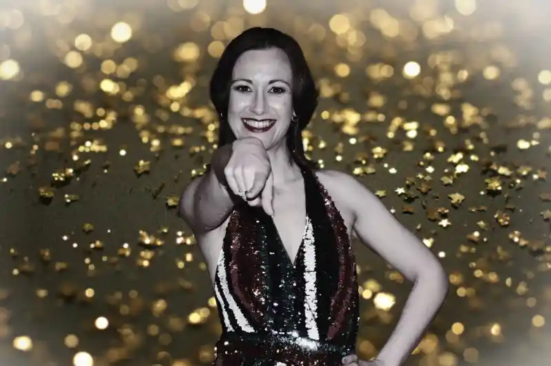 A woman in a sequined dress with bold stripes poses confidently, pointing playfully at the camera. The background features a shimmering golden sparkle effect.