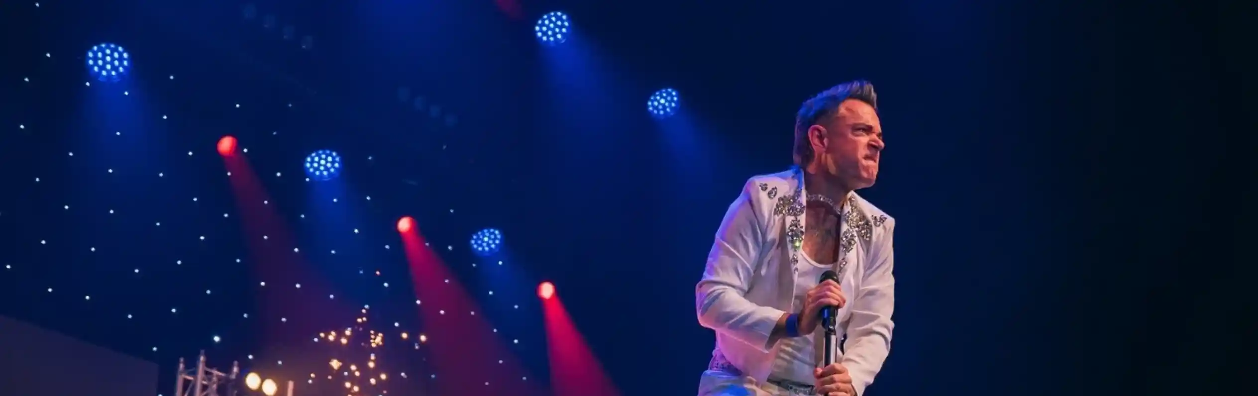 A performer dressed in a white jumpsuit leaps above a microphone stand on stage, with colorful lights and a drum set in the background. The scene captures dynamic energy and excitement during a live music performance.