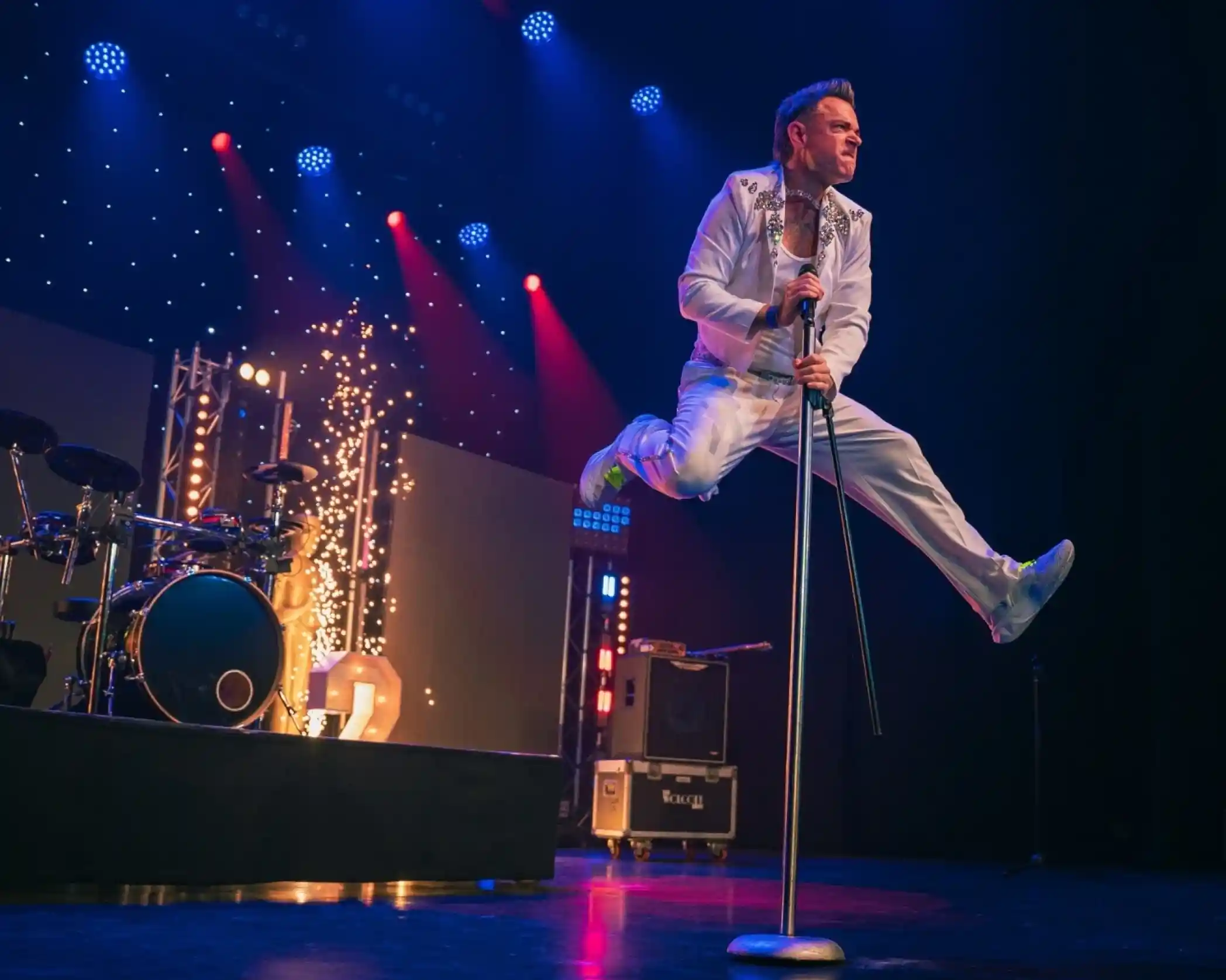 A performer dressed in a white jumpsuit leaps above a microphone stand on stage, with colorful lights and a drum set in the background. The scene captures dynamic energy and excitement during a live music performance.