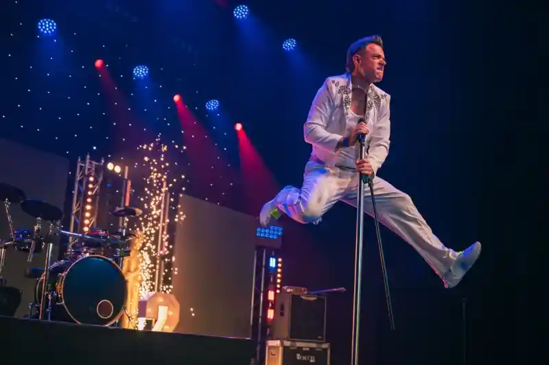 A performer dressed in a white jumpsuit leaps above a microphone stand on stage, with colorful lights and a drum set in the background. The scene captures dynamic energy and excitement during a live music performance.