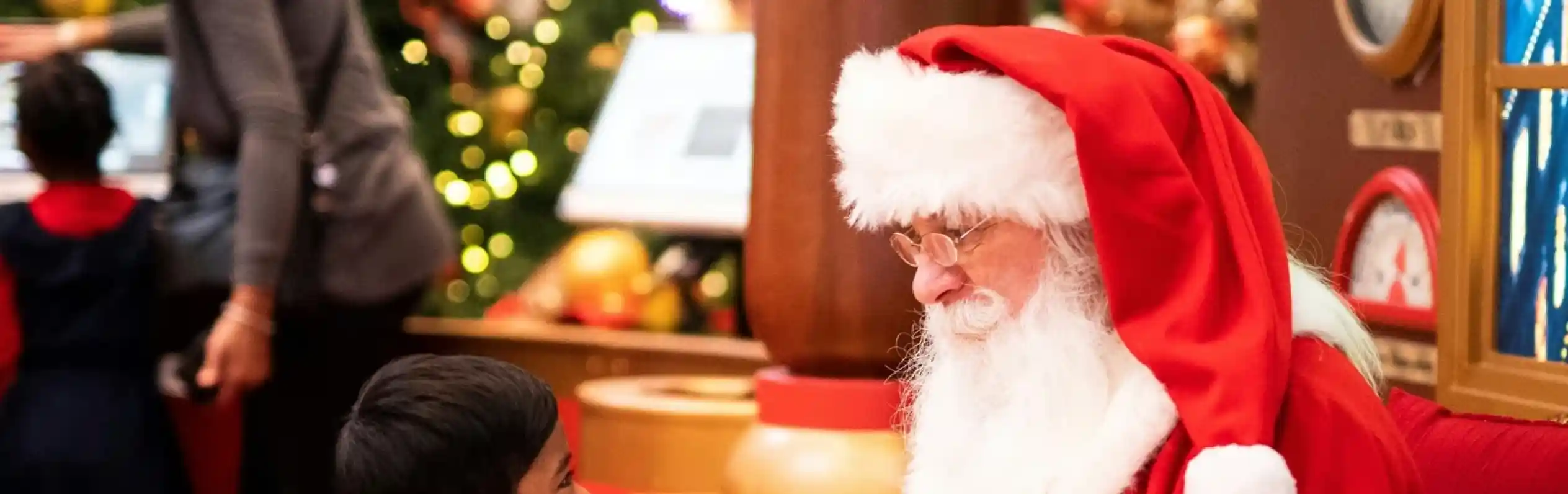 A young boy smiles and leans forward as he talks to Santa Claus, who is seated and dressed in his traditional red suit with white trim. Santa holds a piece of paper and looks attentively at the boy. The background features festive holiday decorations.