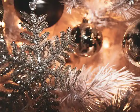 Silver snowflake ornament hangs among shiny golden and black baubles on a decorated white Christmas tree, with warm lights in the background.