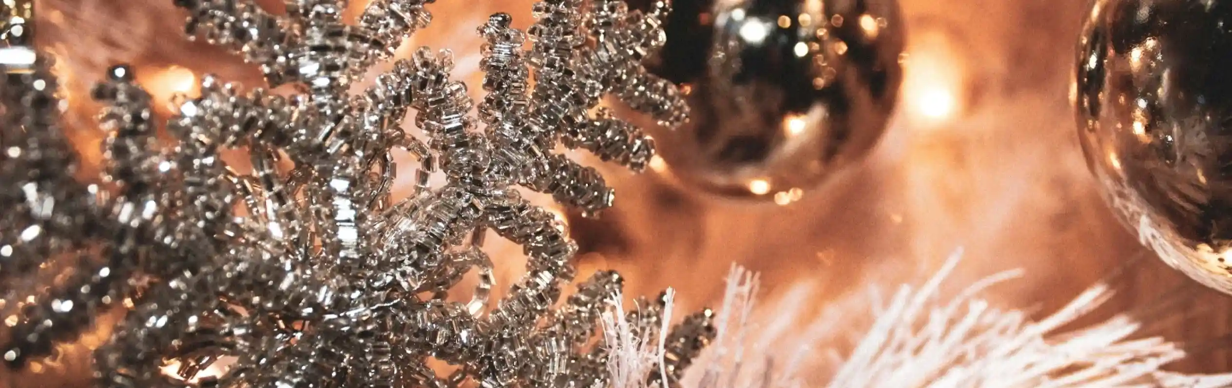 Silver snowflake ornament hangs among shiny golden and black baubles on a decorated white Christmas tree, with warm lights in the background.