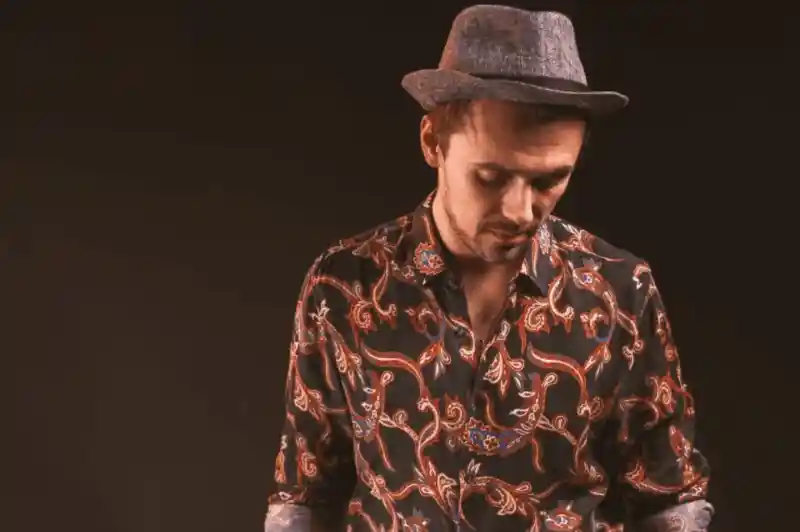 A man wearing a patterned shirt and a grey fedora hat looks down thoughtfully against a dark background.