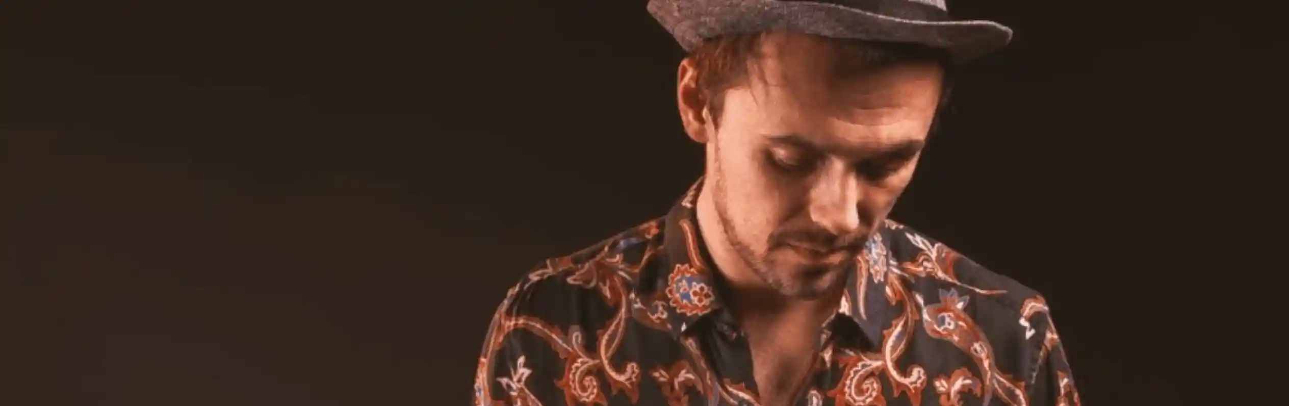 A man wearing a patterned shirt and a grey fedora hat looks down thoughtfully against a dark background.