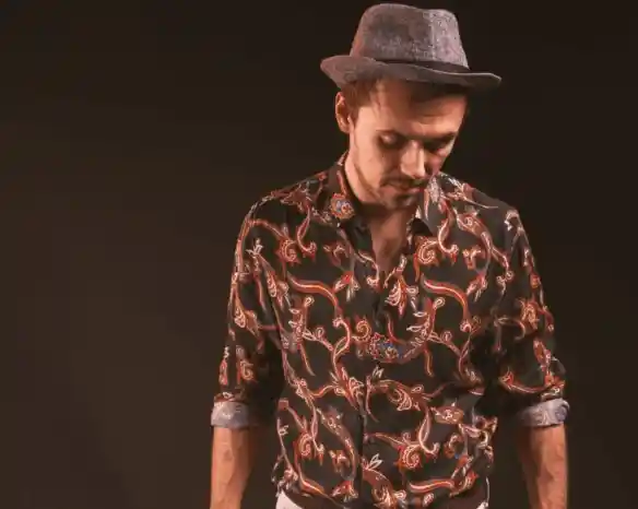 A man wearing a patterned shirt and a grey fedora hat looks down thoughtfully against a dark background.