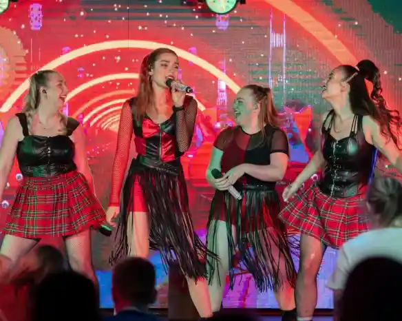 Four performers in matching outfits with fringes and plaid skirts are singing on stage. The background features bright, colorful lights and abstract patterns. The atmosphere is energetic and lively.