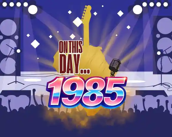 A vibrant graphic featuring a silhouette of a guitar and a microphone, set against a blue background with spotlights. The text reads "On This Day... 1985" in bold, colorful letters, with a crowd silhouette cheering at the bottom.