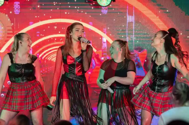 Four performers in matching outfits with fringes and plaid skirts are singing on stage. The background features bright, colorful lights and abstract patterns. The atmosphere is energetic and lively.