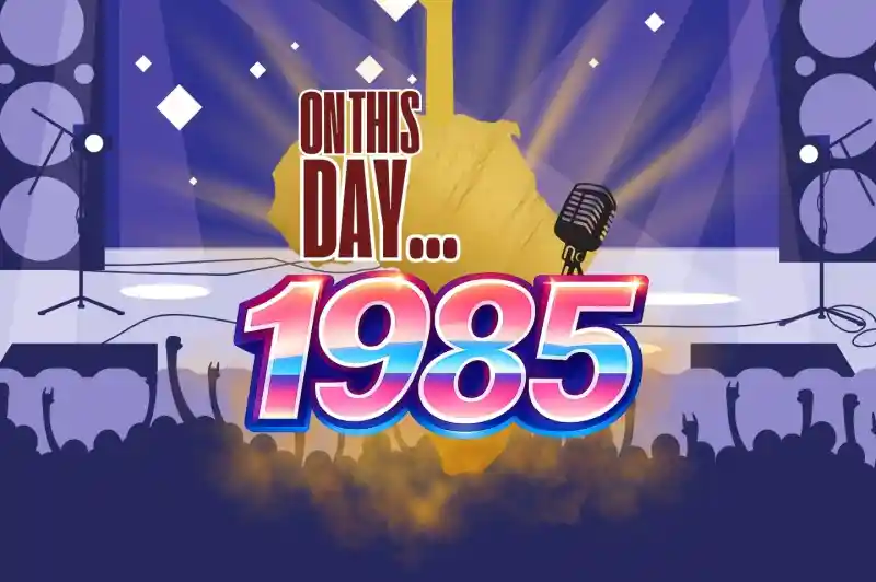 A vibrant graphic featuring a silhouette of a guitar and a microphone, set against a blue background with spotlights. The text reads "On This Day... 1985" in bold, colorful letters, with a crowd silhouette cheering at the bottom.