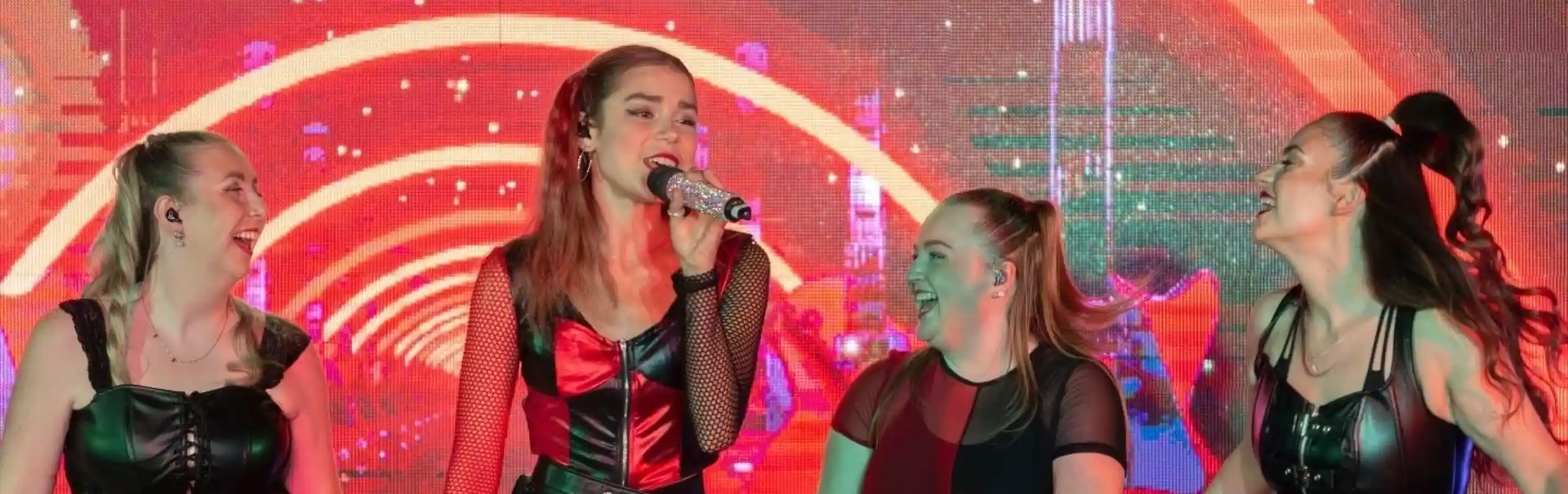 Four performers in matching outfits with fringes and plaid skirts are singing on stage. The background features bright, colorful lights and abstract patterns. The atmosphere is energetic and lively.