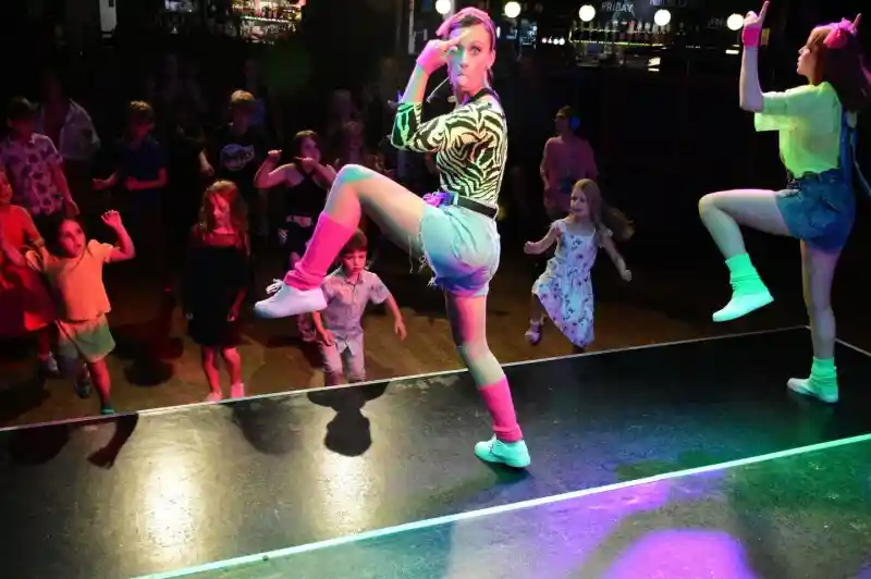 Two performers in colorful outfits dance energetically on stage while a group of children enthusiastically watch and mimic their movements in a lively venue. Bright stage lights create a fun atmosphere.