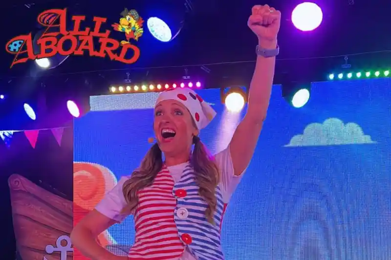A performer dressed in a striped shirt and colorful accessories raises a fist in excitement on stage. Bright lights and a playful backdrop enhance the festive atmosphere.