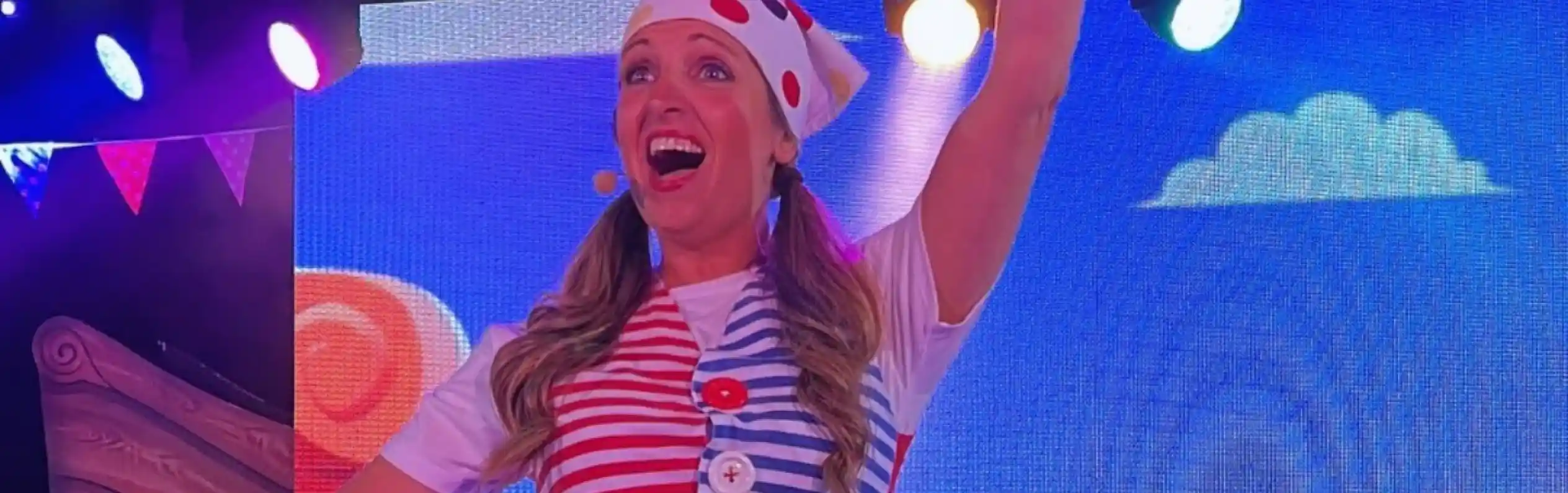 A performer dressed in a striped shirt and colorful accessories raises a fist in excitement on stage. Bright lights and a playful backdrop enhance the festive atmosphere.