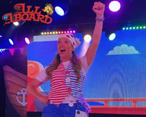 A performer dressed in a striped shirt and colorful accessories raises a fist in excitement on stage. Bright lights and a playful backdrop enhance the festive atmosphere.