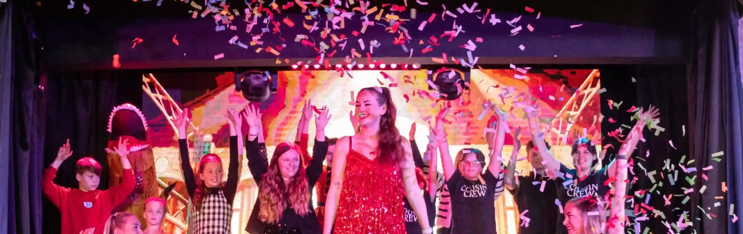 A vibrant stage performance featuring a group of performers celebrating with colorful confetti. The central performer, dressed in a sparkling red dress, strikes a pose, while others in various costumes dance enthusiastically around her.