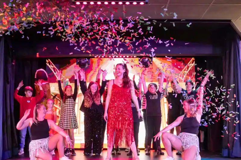 A vibrant stage performance featuring a group of performers celebrating with colorful confetti. The central performer, dressed in a sparkling red dress, strikes a pose, while others in various costumes dance enthusiastically around her.