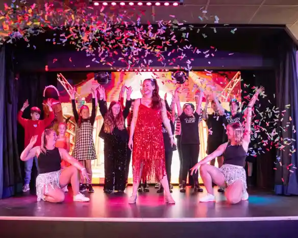 A vibrant stage performance featuring a group of performers celebrating with colorful confetti. The central performer, dressed in a sparkling red dress, strikes a pose, while others in various costumes dance enthusiastically around her.