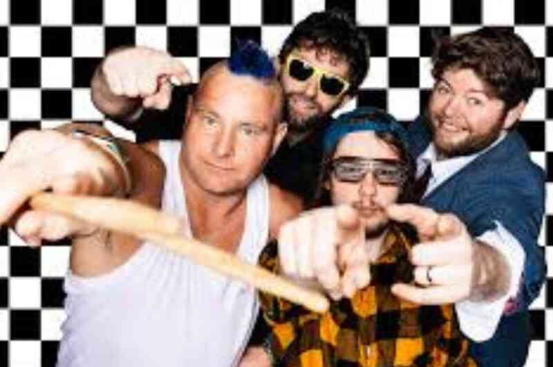 Four men pose in front of a black and white checkered background. One man displays a mohawk hairstyle and is wearing a tank top. Another wears sunglasses and a plaid shirt, while a third sports a blue beanie and sunglasses. The fourth man is dressed in a suit and tie, all are making playful gestures toward the camera.