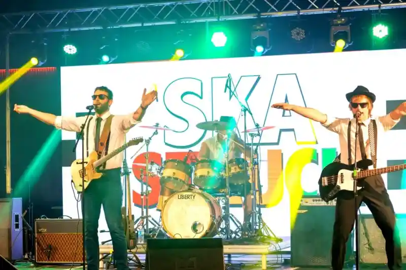 A lively band performs on stage with bright lights and a colorful backdrop. Two musicians, wearing sunglasses and hats, pose with their arms outstretched, while a drummer plays in the background. The atmosphere is energetic and vibrant.