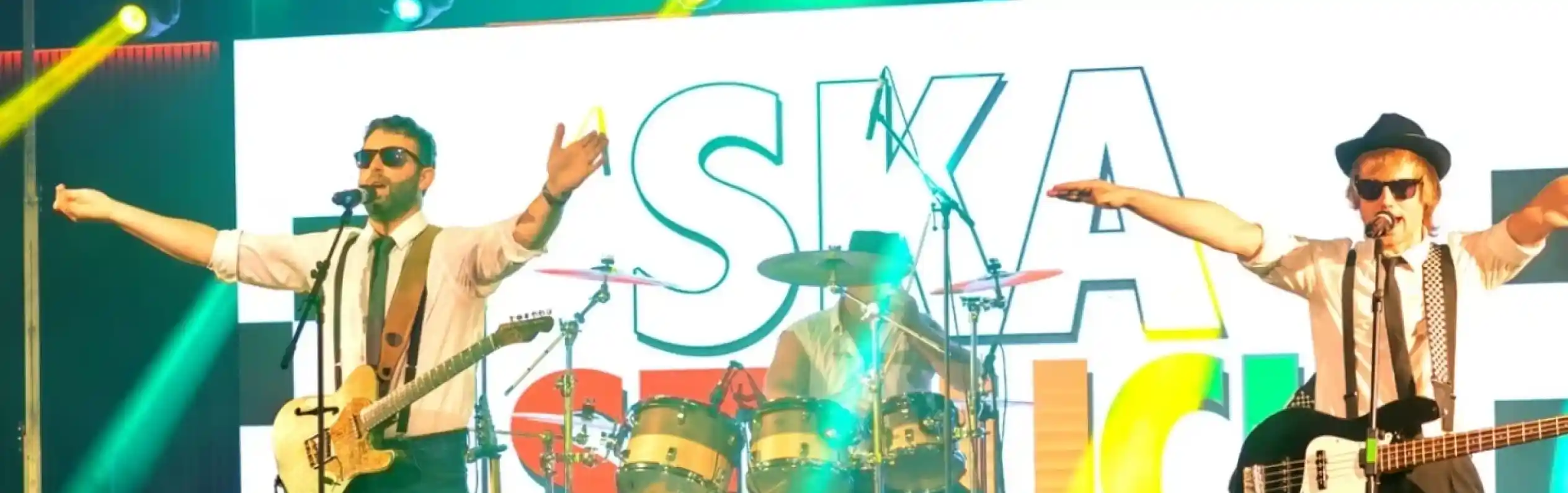 A lively band performs on stage with bright lights and a colorful backdrop. Two musicians, wearing sunglasses and hats, pose with their arms outstretched, while a drummer plays in the background. The atmosphere is energetic and vibrant.
