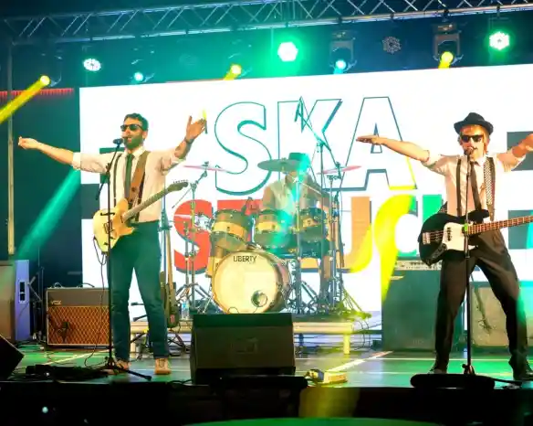 A lively band performs on stage with bright lights and a colorful backdrop. Two musicians, wearing sunglasses and hats, pose with their arms outstretched, while a drummer plays in the background. The atmosphere is energetic and vibrant.