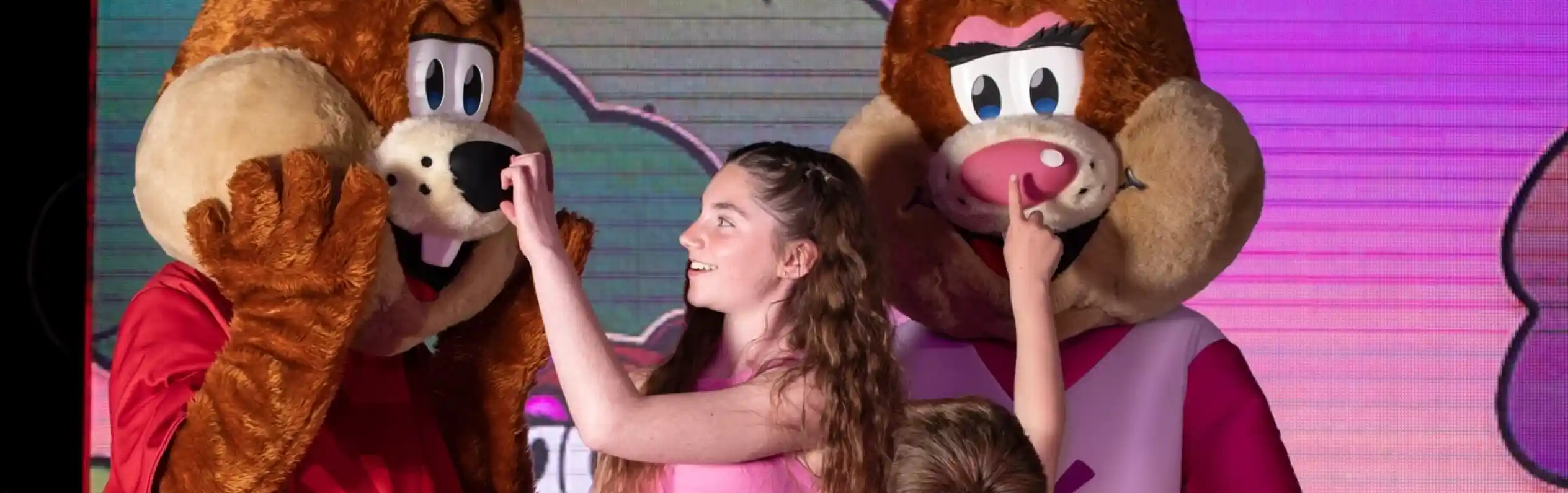 A young girl and boy interacting playfully with two costumed performers acting as squirrel characters on stage. Another performer holds a puppet dog next to them.