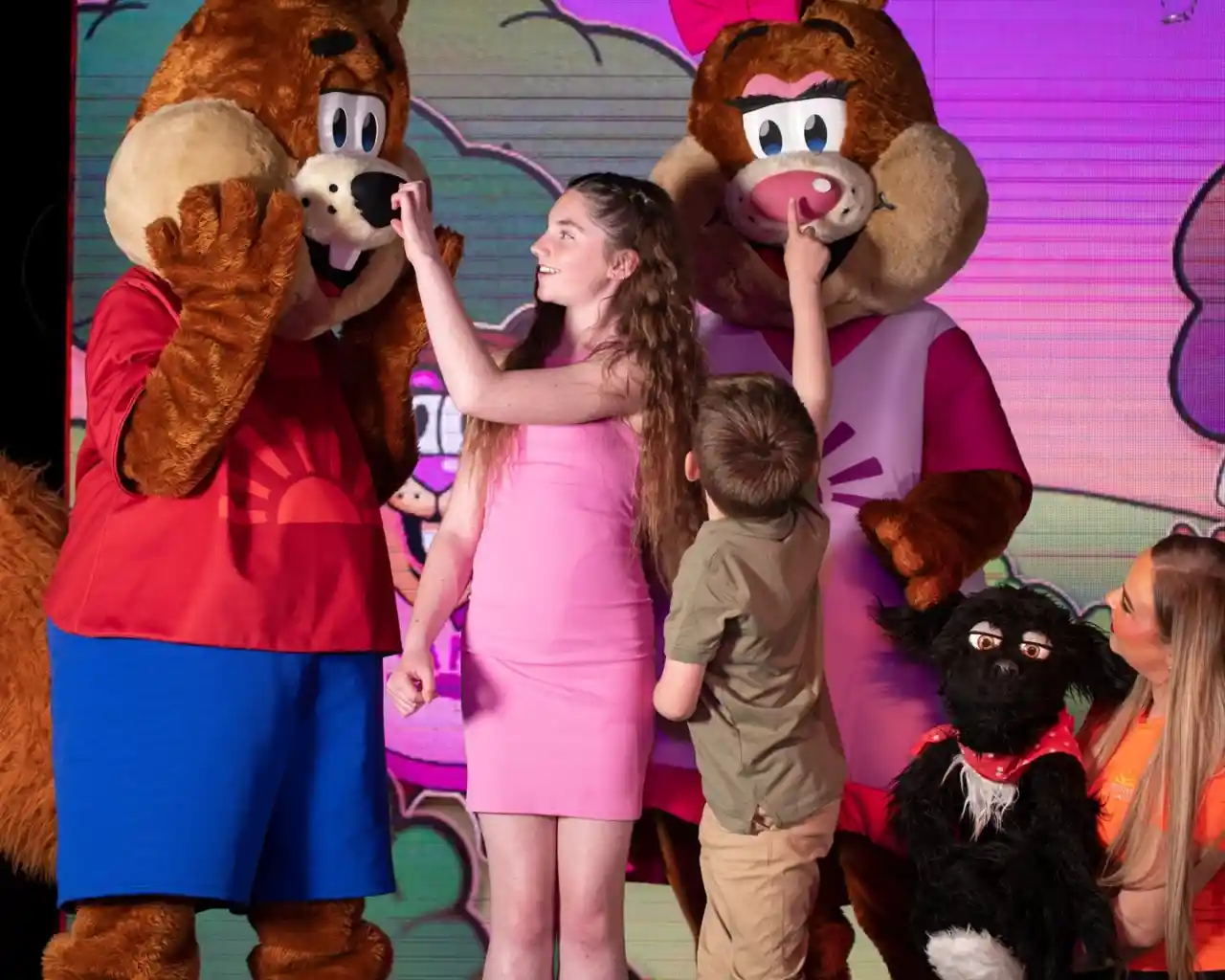 A young girl and boy interacting playfully with two costumed performers acting as squirrel characters on stage. Another performer holds a puppet dog next to them.