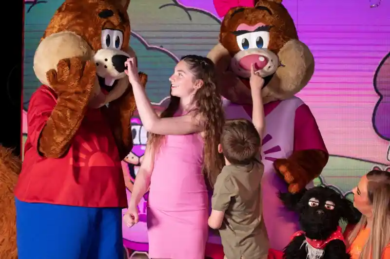 A young girl and boy interacting playfully with two costumed performers acting as squirrel characters on stage. Another performer holds a puppet dog next to them.
