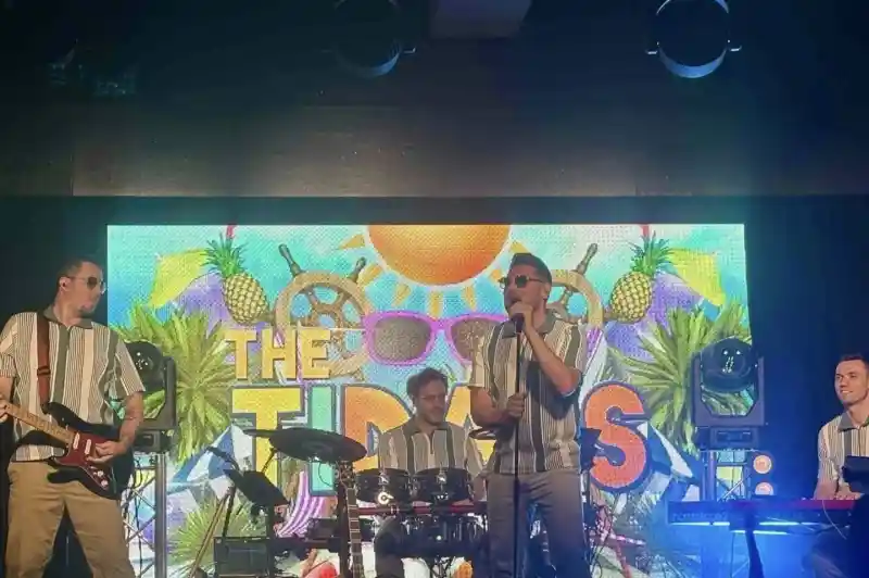A lively band performs on stage, dressed in matching striped shirts and beige pants. The setting features a vibrant backdrop with tropical elements like pineapples and sunglasses, along with bright stage lights. The musicians include a vocalist with sunglasses, a guitarist, a drummer, and a keyboardist, creating an energetic atmosphere.
