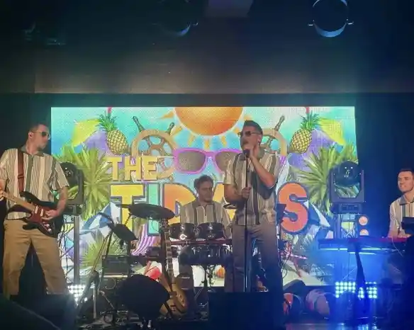 A lively band performs on stage, dressed in matching striped shirts and beige pants. The setting features a vibrant backdrop with tropical elements like pineapples and sunglasses, along with bright stage lights. The musicians include a vocalist with sunglasses, a guitarist, a drummer, and a keyboardist, creating an energetic atmosphere.