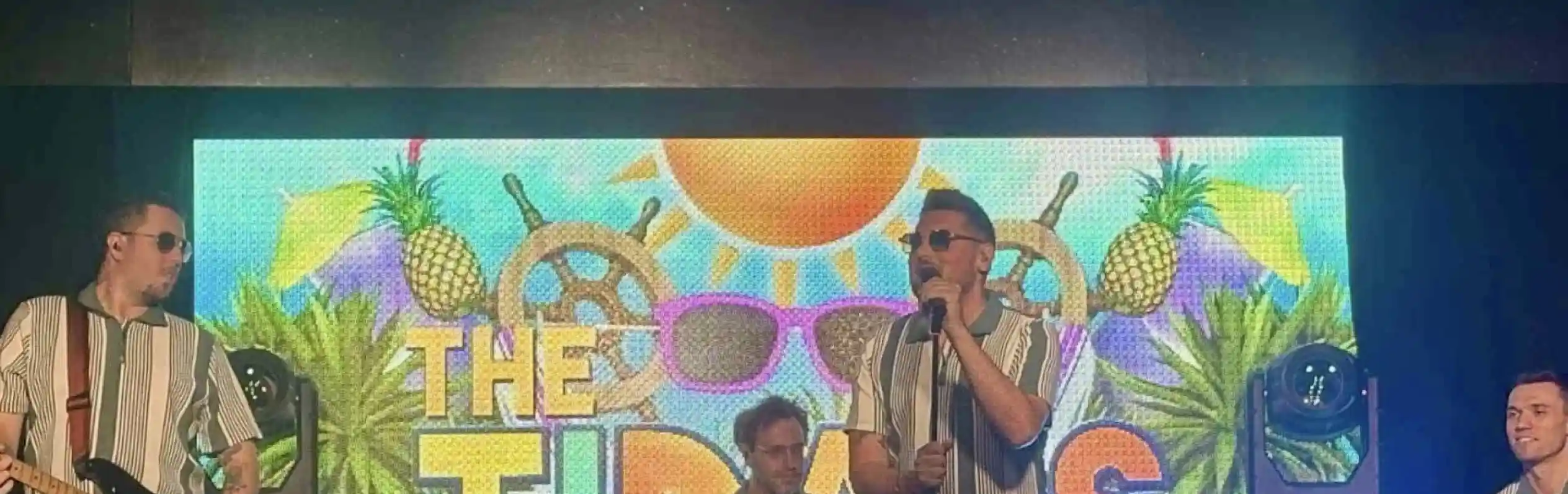 A lively band performs on stage, dressed in matching striped shirts and beige pants. The setting features a vibrant backdrop with tropical elements like pineapples and sunglasses, along with bright stage lights. The musicians include a vocalist with sunglasses, a guitarist, a drummer, and a keyboardist, creating an energetic atmosphere.