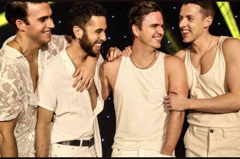 Four young men laugh together on stage, dressed in light-colored outfits. They stand close, showcasing camaraderie and joy, with colorful stage lights glowing in the background.