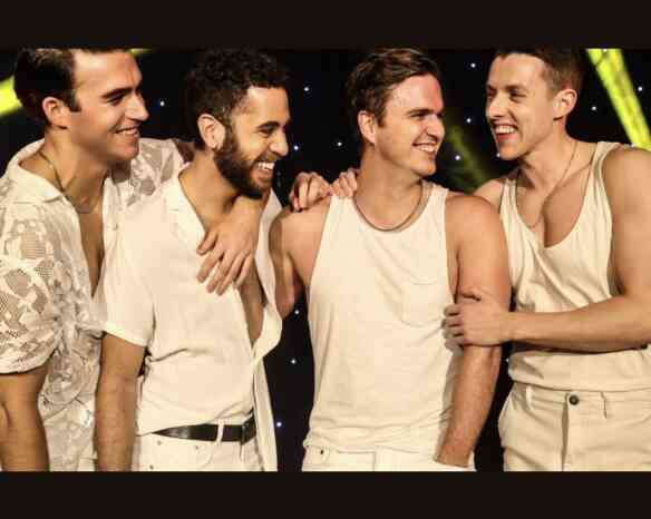 Four young men laugh together on stage, dressed in light-colored outfits. They stand close, showcasing camaraderie and joy, with colorful stage lights glowing in the background.