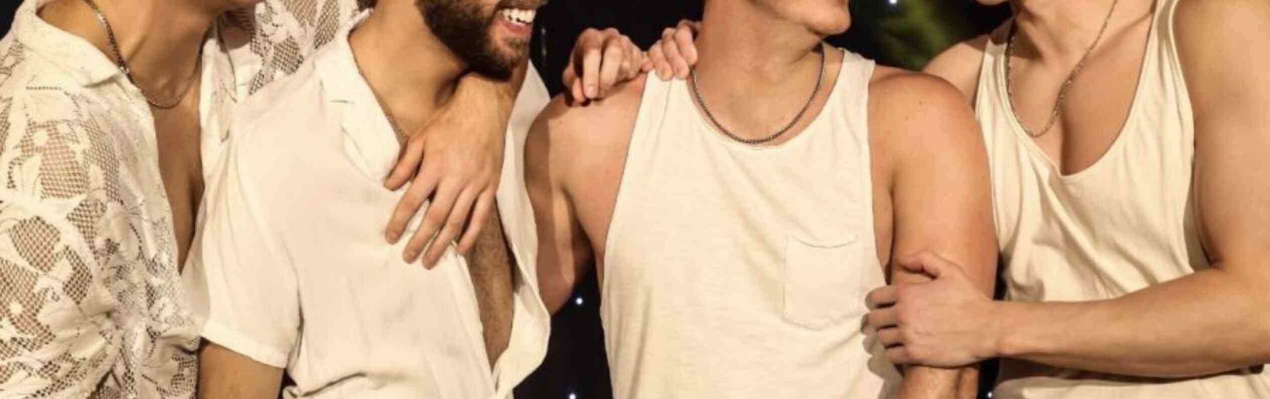 Four young men laugh together on stage, dressed in light-colored outfits. They stand close, showcasing camaraderie and joy, with colorful stage lights glowing in the background.