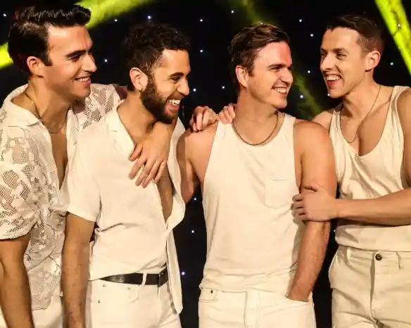 Four men dressed in casual white outfits laugh and interact with each other on stage, illuminated by colorful lights. They display camaraderie and joy as they smile at one another.